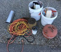 Overview photo showing vintage gas can, two buckets of tools, electrical cords, and rolls of materials arranged on gravel surface.