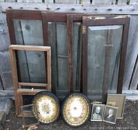Photo showing the set of four large wooden window frames including three with beveled glass, two oval decorative mirrors, two black and white photographic portrait frames, one small mesh-fronted frame and one small wooden frame without glass, all leaning against a wooden fence.