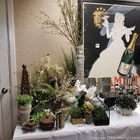 Wide shot showing a variety of vase types with faux plants, decorative metal and ceramic objects on a table covered with a white cloth.