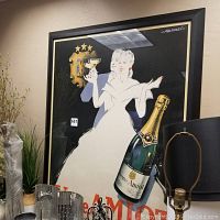 Full view of framed artwork showing a woman in white holding a glass and a large Veuve Amiot champagne bottle, frame black with some dents and paint spots visible