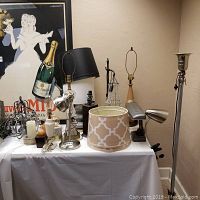 Photo showing a variety of lamps with bases in metal, ceramic, and wood, several fabric lampshades, and assorted candleholders and candles on a table against a wall.