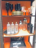 Bar set displayed inside wooden cabinet with three shelves. Top shelf holds black and gold rimmed glasses and plates. Middle shelf holds crystal bar shakers, labeled liquor bottles on brass and black leather tray, and a black case. Bottom shelf has Travel Bar boxed travel set with metal cups in black leather case.