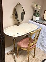Wide angle of vintage vanity table with marble top, gold designs, gold legs and attached oval mirror. Gold-painted chair with pink cushion in front.