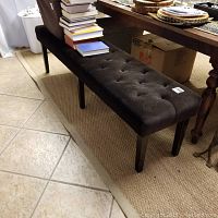 Long tufted upholstered bench with dark fabric showing wear and button details, placed on a tan rug on tiled floor.