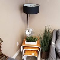 Overall view showing three wooden nesting tables stacked, standing floor lamp behind, metal fan placed on top, faux green plant in white planter on table.