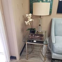 Full view of glass top side table with metal legs, vintage lamp on top, faux orchid plant, and small wooden tray.