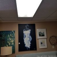 Canvas print signed 'Alm' with stylized white figure on black background, flanked by two watercolor paintings in white frames on wall.