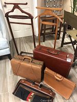 Photo showing two wooden clothes valets behind four pieces of luggage: a red suitcase, camel leather briefcase, small tan briefcase, and canvas leather travel bag.