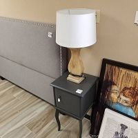 Dark grey wooden side table with lamp on top, next to a grey bed frame with nailhead trim and framed artwork leaning on the wall.