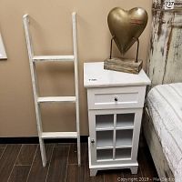 View of white wood cabinet with marble top next to white distressed wooden ladder and gold-toned metal heart decor on wooden base.