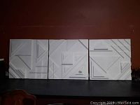 Set of three white square geometric relief art pieces arranged side by side showing abstract angular designs
