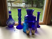 Lot items arranged on a windowsill showing all from left to right: cut glass decanter, candlesticks, glass bottles, vase, and shot glasses in front