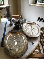 Photo of the full lot on a round table showing silverplate serving platters with leaf designs, gravy boat, pewter pitcher, and vase.