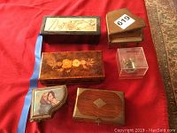 Photo showing six vintage music boxes arranged on a carpet, including rectangular wood boxes with inlaid flowers, a piano-shaped box, an acrylic case, and a piano box with a Madonna cloth picture.