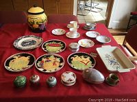 All lot contents arranged on red tablecloth, including decorative plates, trinket boxes, cups, saucers, and the biscotti jar.