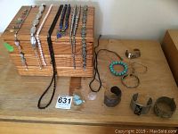 Image shows multiple necklaces and bracelets arranged on and in front of a wooden jewelry box. Includes black bead necklace, faux pearl necklaces, turquoise bead bracelet, stone pendant necklace, quartz earrings, and several metal bracelets, one large Mexican silver cuff bracelet.