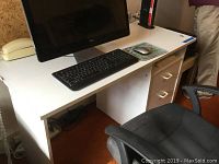 White laminate computer desk with computer monitor, keyboard, and mouse on top (items not included). Desk shows wear and damage especially on front left edge.