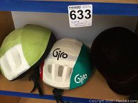 Two bicycle riding helmets, one green and white with mesh, and one Giro helmet in white and green with black straps on shelf with auction sticker.