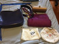 Five vintage purses including a purple leather shoulder bag (Italy) with stain, metallic gold hard clutch (France), maroon clutch, cream woven clutch, and small floral embroidered purse.