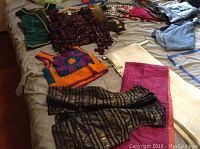 Photo of multiple clothing items including green velvet vest with gold trim, black and gold sheer vest, hand-knitted multicolored vest, scarves, poncho, and linens laid out on bed.