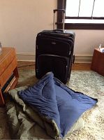 Black 29 inch rolling luggage and folded cold weather sleeping bag on green carpet near wooden furniture.
