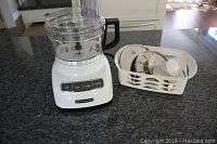 White KitchenAid food processor with control buttons on granite countertop and basket with discs beside it.
