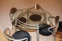 Clear image showing round beveled glass table with metal base and four metal chairs with black upholstered seats arranged around it.