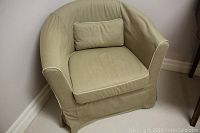 Front side angle of beige upholstered tub chair with lumbar pillow and white piping on cushions.