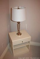 Side table with drawer and two shelves, lamp on top, showing table and lamp in corner