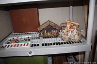 Two nativity scenes on a shelf, one traditional stable with figurines and one colorful musical ceramic set, along with a container of small figures.