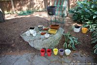 Multiple flower pots and small planters placed on and around a large rock outdoors; showing a variety of colors, shapes, and sizes.