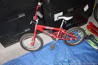 Full side view of the red Norco Macho kids bicycle showing frame, saddle, pedals, training wheels, and wheels on a blue tarp with a black background.