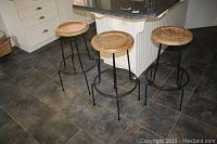 Three metal bar stools with round wooden seats placed in a kitchen setting on tiled floor.
