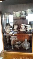 Wooden cabinet displaying bone china teacups, saucers, glass champagne flutes, and cut glass covered dish.