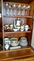 Wooden shelving unit with 7 wine glasses on top shelf, Royal Albert china teapot, sugar, creamer and cups on middle shelf, and variety of plates and cups on bottom shelf