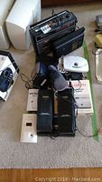 Wider view of all electronics included in the lot: Transonic cassette player radio with answer machine, two black Realistic Citizen Band transceivers, Cosmo radio device, headphones, manuals, and cables.