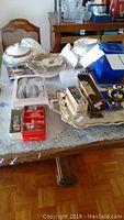 Wide view of table showing silver plated tray, cake plates, napkin rings, boxed utensils, crystal goblets, and casserole set.