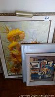 Photo showing stacked framed artworks leaning against a wall, largest floral floral painting in gold-toned frame visible on top