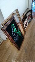 Several framed art pieces and an ornate mirror leaning against a wall on wooden floor