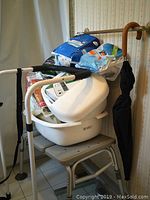 Photo showing a white plastic tub bench with attached handles, a beige/grey foldable bed grip, multiple unopened packages of adult diapers stacked on top, and a black folding umbrella leaning against a wall.