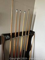 Five wooden pool cue sticks placed upright in a dark wood wall rack with five slots intended for holding cues.