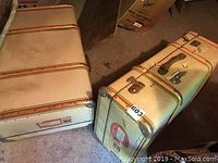 Two vintage beige suitcases stacked on floor with metal trims and leather straps.