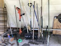 Full view of garden tools including various rakes, shovels, and broom next to a wall with extension cord on the ground