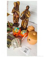 Photo of Mexican collectible figurines, pottery, gourds, wooden dishes, mortar and pestle, and a colorful wind instrument arranged on a white surface.