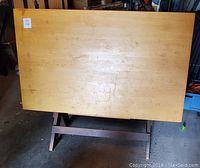 Rectangular wooden drafting table surface showing wear and marks, resting on angled wooden base.