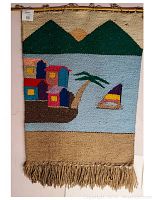 Handmade Mexican hanging blanket with colorful scenic woven design showing houses by a lake, sailboat, green mountains, and a sun. Finished with fringe at the bottom.