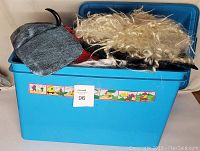 A blue plastic trunk containing various wigs, masks, hats, and costume pieces including a blonde curly wig and a grey fabric hat.
