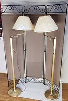 Two brass adjustable floor lamps with cream pleated lamp shades set in front of three-panel black metal framed bi-fold screen with fabric inserts and decorative scrollwork.