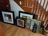 Eight framed art pieces leaning against a wall near stairs, showing varied abstract and figurative artwork in different frame styles and sizes.