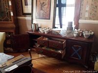Wood cabinet with cups, teacups stacked, punch bowl, ice bucket, various dishes wrapped in newspaper inside and on top; some drawers open showing more items.